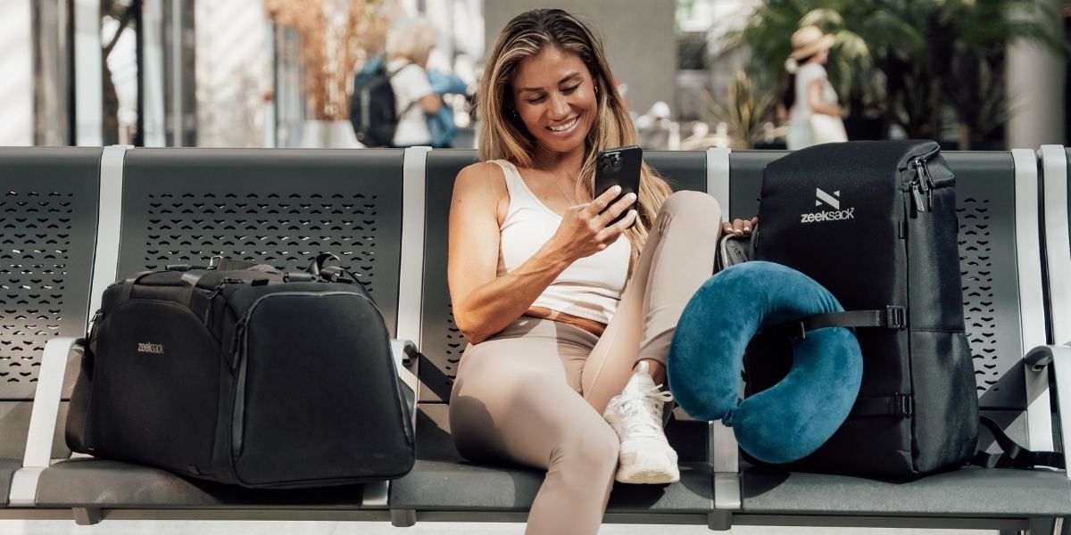 Frau sitzt am Flughafen und schaut auf ihr Handy, neben ihr steht ein schwarzer Zeeksack Reiserucksack mit Nackenkissen und eine Reisetasche, ideales Handgepäck für Flugreisen.