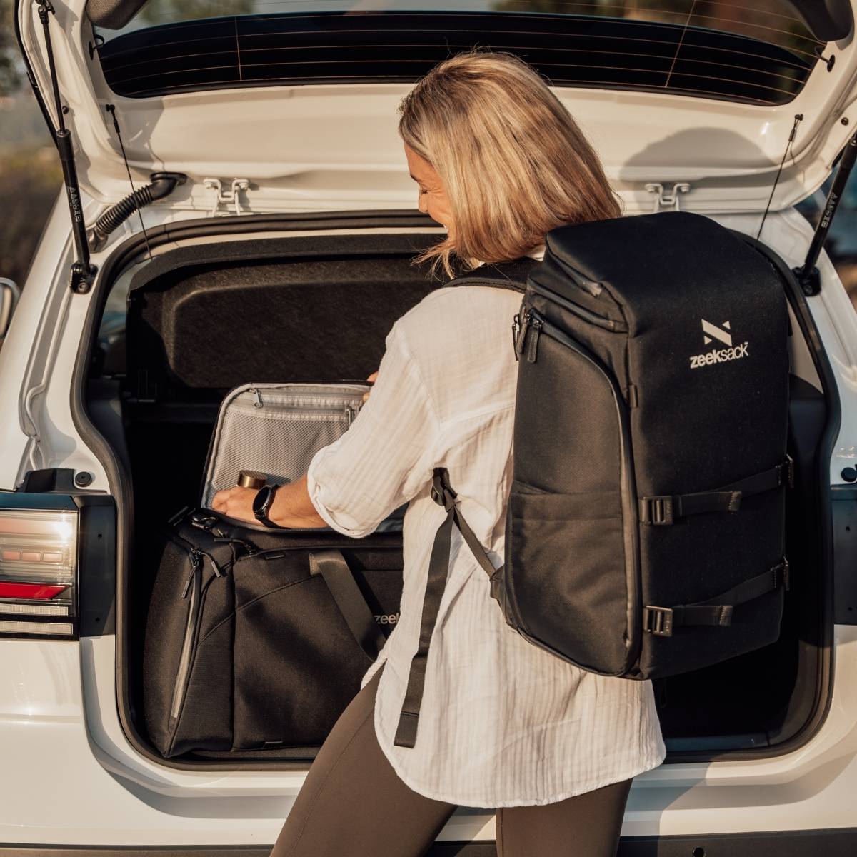 Frau mit schwarzem Rucksack vor einer geöffneten Kofferraumklappe.