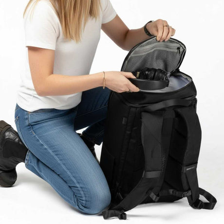 Frau kniet und legt Kopfhörer in das obere Fach eines schwarzen Zeeksack Rucksacks, praktischer Rucksack mit organisiertem Stauraum für Alltag und Reisen.